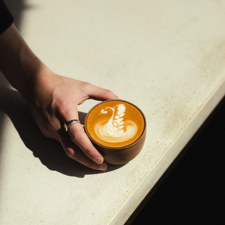 A flat white coffee in a cup, held by a hand over a table, with soft shadows cast by natural light.